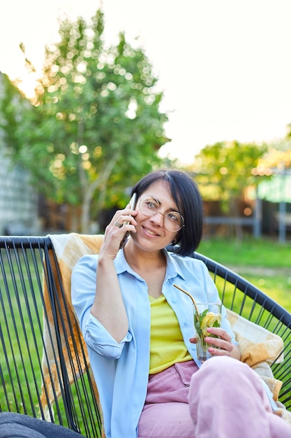 Szczęśliwa kobieta w okularach rozmawiająca przez telefon komórkowy siedząca na krześle na podwórku na świeżym powietrzu