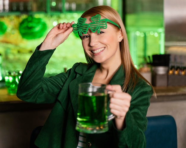 Zdjęcie szczęśliwa kobieta w okularach shamrock świętuje ul. patrick's day z drinkiem w barze