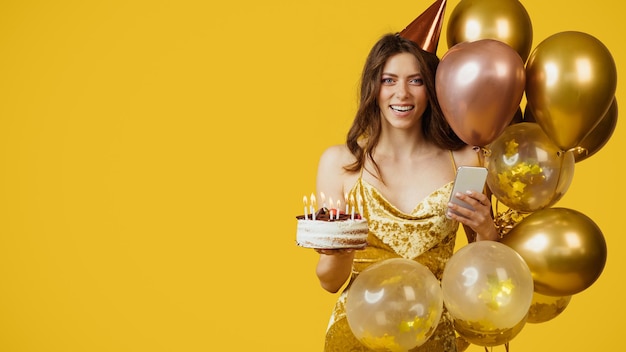 Szczęśliwa kobieta w sukience i kapeluszu urodzinowym trzymająca tort ze świeczkami i używająca telefonu komórkowego stojącego z balonami wolnej przestrzeni