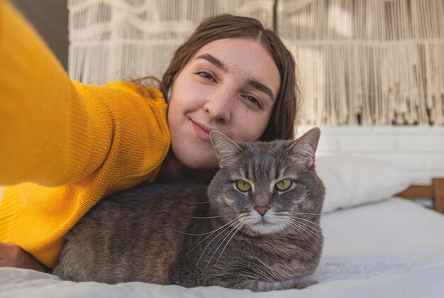 Szczęśliwa kobieta w żółtym swetrze przytula swojego szarego kota na łóżku w lekkim wnętrzu