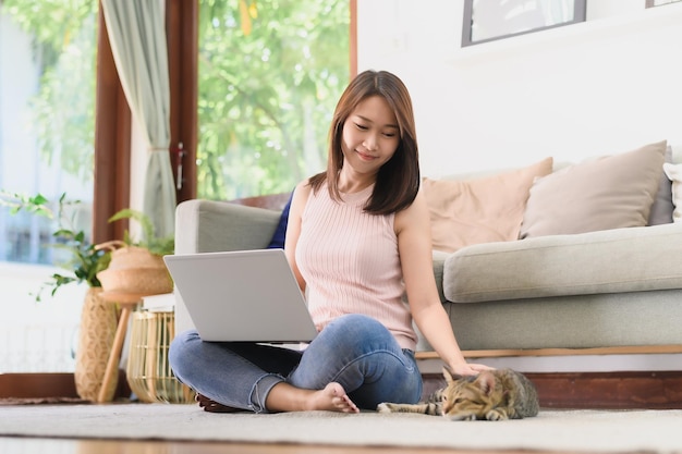 Szczęśliwa kobieta z Azji bawi się z kotem podczas korzystania z laptopa w salonie Praca w domu koncepcja