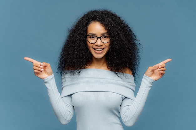 Szczęśliwa kobieta z fryzurą Afro, wskazuje na boki dwoma palcami wskazującymi, nosi swobodny sweter