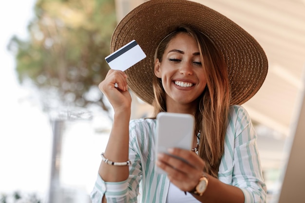 Szczęśliwa kobieta z kartą kredytową, korzystająca z telefonu komórkowego i sprawdzająca swoje konto bankowe podczas relaksu w kawiarni