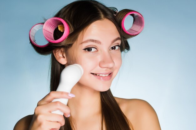 Zdjęcie szczęśliwa kobieta z lokówkami na głowie czyści skórę twarzy szczoteczką do głębokiego czyszczenia