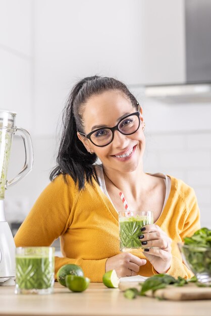 Szczęśliwa kobieta ze szpinakowym smoothie lub wegetariańskim shake'iem siedząca w swojej kuchni