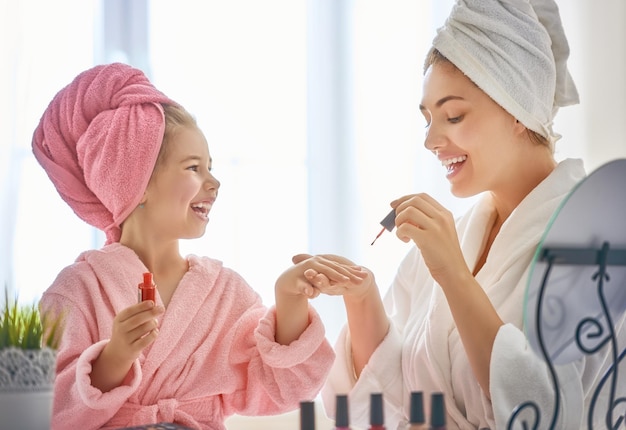 Szczęśliwa kochająca rodzina Matka i córka robią manicure i bawią się siedząc przy toaletce w domu Mama i dziecko dziewczynka są w szlafrokach i ręcznikach na głowach