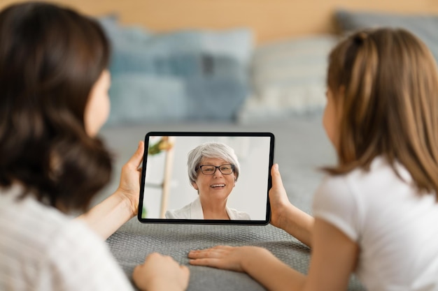 Szczęśliwa kochająca rodzina. Młoda matka i córka dziewczyna przy użyciu komputera typu tablet do zdalnej rozmowy z babcią. Zabawna mama i kochane dziecko bawią się w domu.