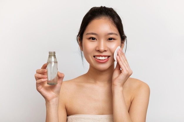 Szczęśliwa koreańska dama trzymająca wacik i butelkę z żelem do mycia twarzy stojące nad białym studio