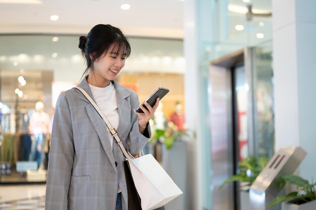 Szczęśliwa, ładna Azjatka sprawdza wiadomości na telefonie podczas spaceru po centrum handlowym