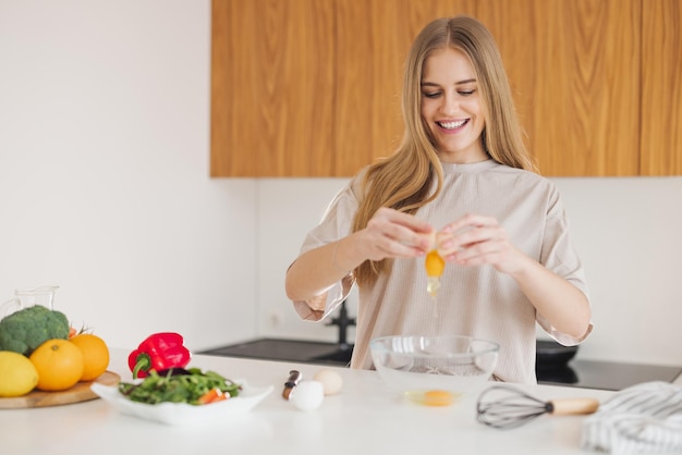 Szczęśliwa ładna blondynka w piżamie przygotowuje śniadanie z jajek i świeżych warzyw w kuchni w domu