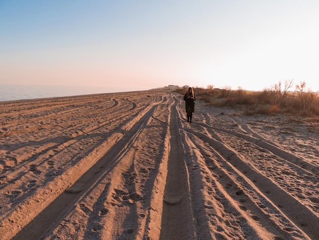 Szczęśliwa ładna młoda kobieta w czarnej kurtce ze smartfonem pusta plaża wieczorem morze zachód słońca Tysiącletnia dziewczyna z długimi włosami