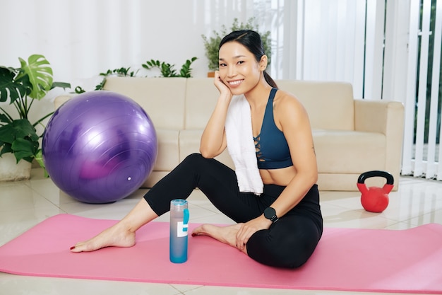 Szczęśliwa ładna młoda wysportowana Azjatka z ręcznikiem na ramieniu spoczywająca na macie do jogi po treningu w domu