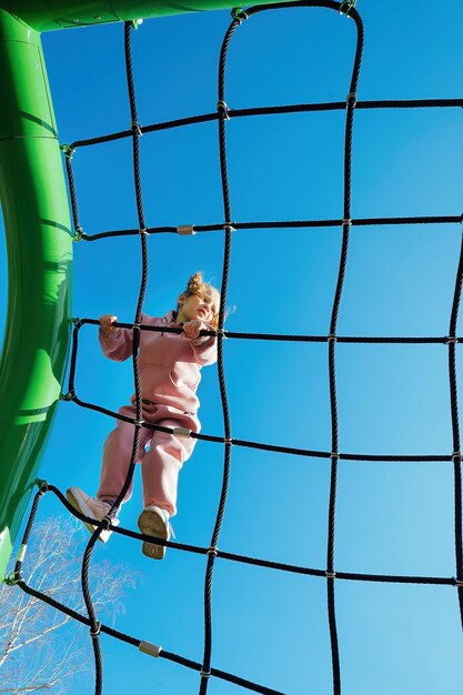 Zdjęcie szczęśliwa, mała, aktywna dziewczyna wspięła się na sieć linową na placu zabaw przed jasnym błękitnym niebem