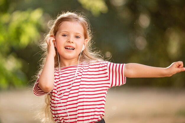 Szczęśliwa mała blondynki dziewczyna słucha muzyka z hełmofonami, tanczy i śpiewa w naturze w parku.