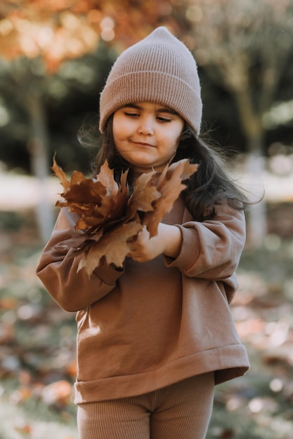 szczęśliwa mała brunetka w czapce z dzianiny spaceruje po parku jesienią