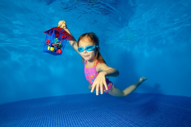 Szczęśliwa mała dziewczynka pływająca pod wodą w basenie z prezentem w ręku na niebieskim tle