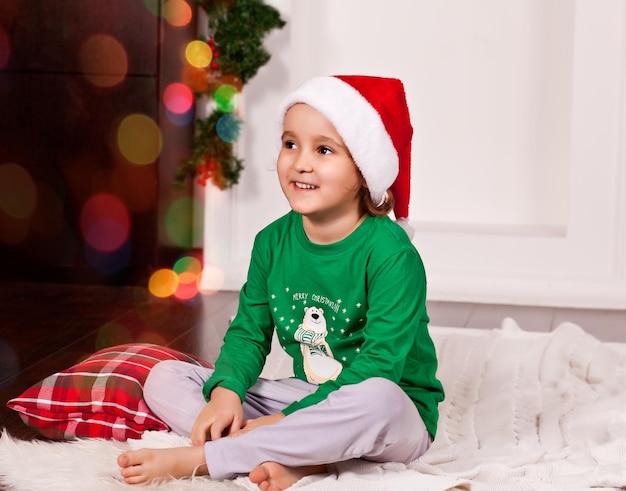 Szczęśliwa mała uśmiechnięta dziewczynka w czapce Mikołaja siedzi na kocu w Boże Narodzenie piżamie.