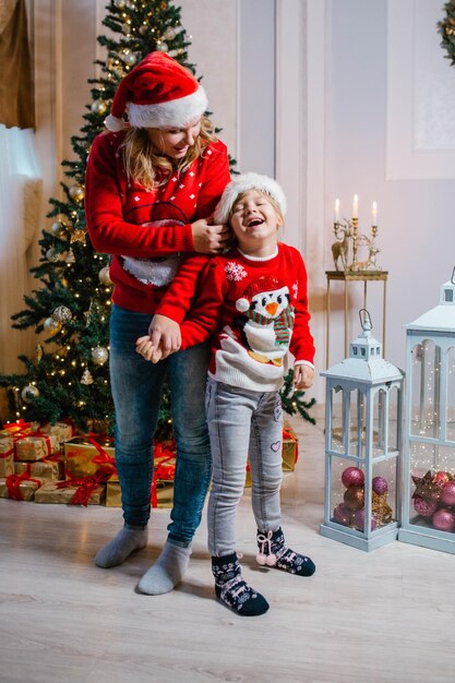 Szczęśliwa mama i śmiejąca się córka w świątecznych swetrach stoją przy choince i bawią się.