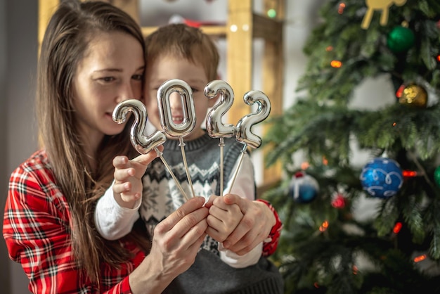 Szczęśliwa mama i syn na tle choinki trzymają w rękach srebrne cyfry 2022 na patykach. Koncepcja Nowego Roku, świąteczna atmosfera, dekoracja domu, gratulacje