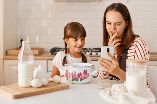 Szczęśliwa mama piecze razem z córką, siedzi przy stole w kuchni i wyszukuje w internecie przepis przez telefon komórkowy, wybiera ciasto, które chce upiec.