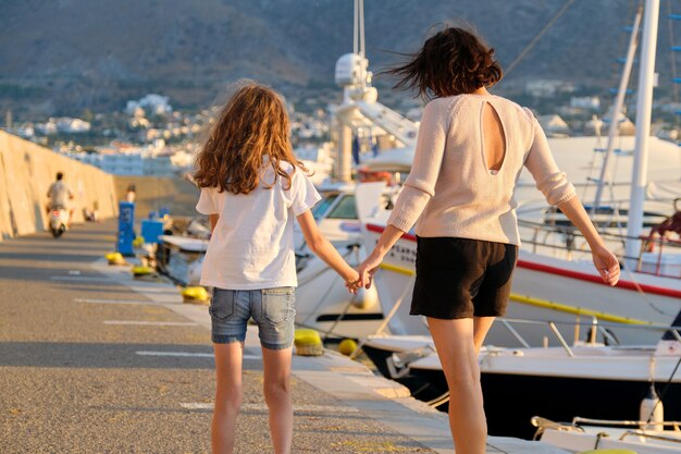 Szczęśliwa matka i córka dziecko spaceru razem trzymając się za ręce wzdłuż morza. Marina dla jachtów, zachód słońca na morzu, tle malowniczych gór. Rodzina, miłość, podróże, styl życia, koncepcja wakacji