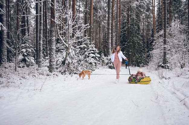 Szczęśliwa matka i mała urocza dziewczynka w różowym ciepłym stroju wierzchnim, chodzenie, zabawa jeździ nadmuchiwaną rurą śnieżną z czerwonym psem shiba inu w śnieżnobiałym zimnym lesie na zewnątrz