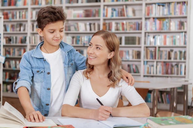 Szczęśliwa matka i syn uśmiechają się do siebie podczas odrabiania lekcji w bibliotece