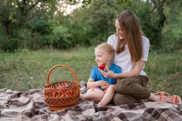 Szczęśliwa matka oferuje jabłko córki blond z kosza siedzącego na kocu w lesie Kobieta z dzieckiem miała piknik w parku miejskim
