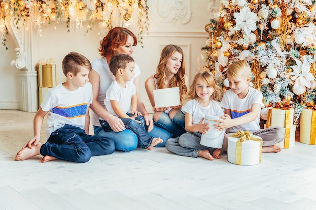 Szczęśliwa Matka Rodziny I Pięcioro Dzieci Relaksuje Się, Bawiąc Się W Pobliżu Choinki W Wigilię Bożego Narodzenia W Domu. Mama, Córki, Synowie W Jasnym Pokoju Z Zimową Dekoracją. Boże Narodzenie Nowy Rok Czas Na świętowanie