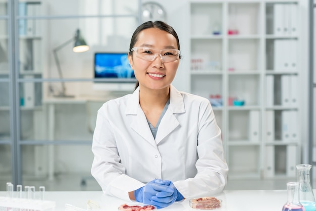 Szczęśliwa młoda Azjatka w rękawiczce w białym fartuchu i okularach siedząca przy miejscu pracy z dwiema próbkami surowego mięsa warzywnego na płytkach Petriego