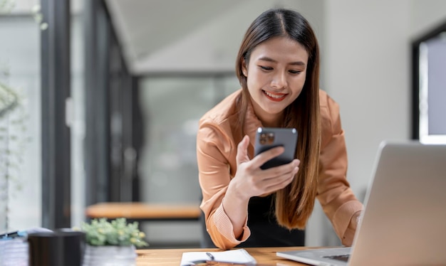 Szczęśliwa młoda azjatycka bizneswoman używająca telefonu komórkowego stojącego przy biurku z laptopem
