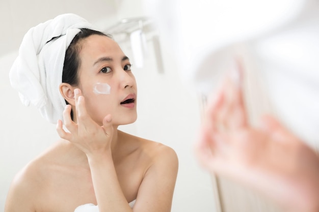 Szczęśliwa młoda azjatycka kobieta stosująca balsamy do twarzy, mając na sobie ręcznik i dotykając twarzy w łazience