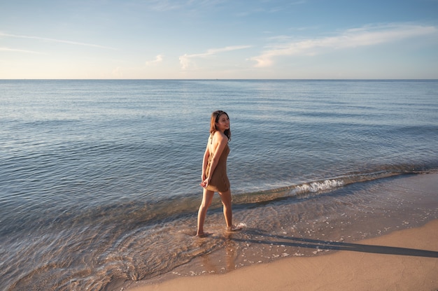 Szczęśliwa młoda azjatycka kobieta w brązowej sukience spacerująca po plaży rano nad tropikalnym morzem na letnie wakacje