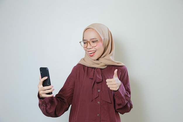 Szczęśliwa młoda azjatycka muzułmańska kobieta w okularach za pomocą telefonu komórkowego odizolowanego na białym tle