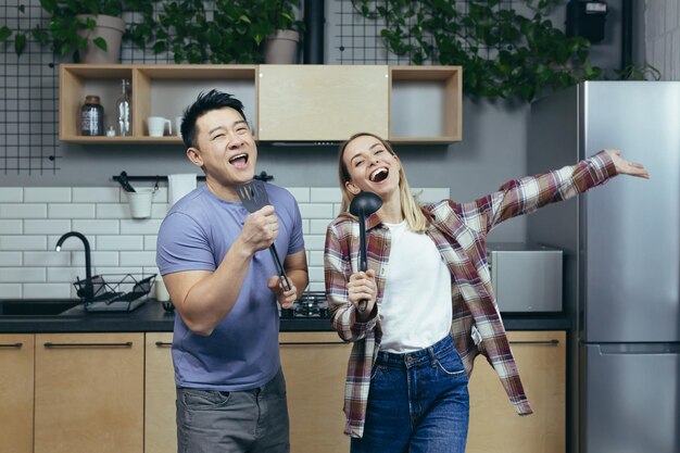 Szczęśliwa młoda azjatycka para mężczyzna i kobieta śpiewa i tańczy razem w kuchni, bawiąc się razem, zakochana rodzina