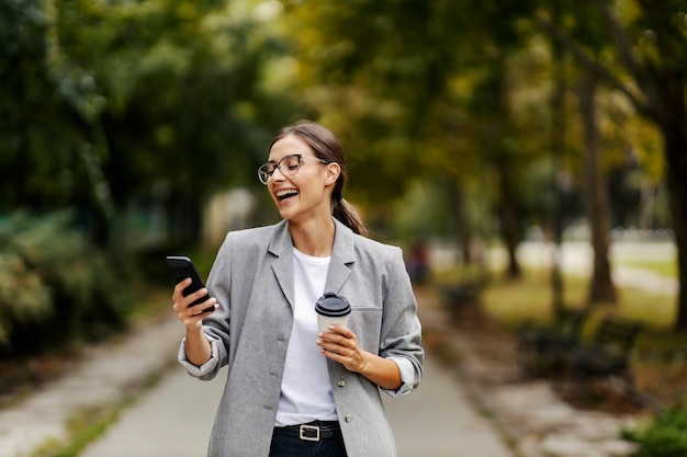 Szczęśliwa młoda bizneswoman śmieje się i trzyma filiżankę kawy, patrząc na telefon