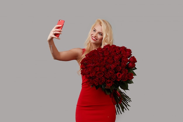 Szczęśliwa Młoda Blond Kobieta Trzyma Duży Bukiet Czerwonych Róż Jako Prezent Na 8 Marca Lub Walentynki. Robi Selfie Na Telefonie Komórkowym