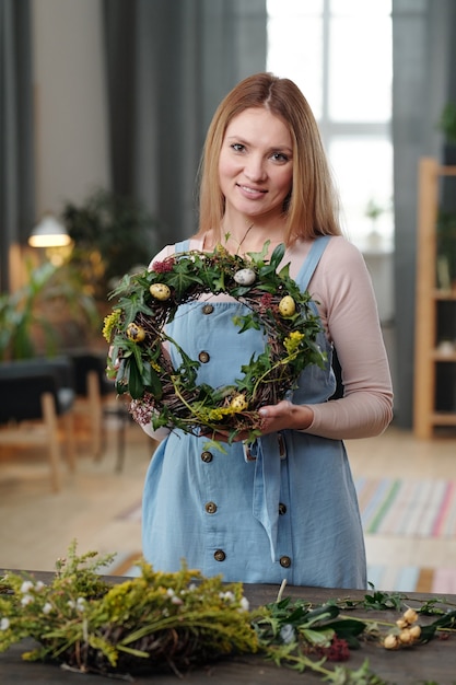 Szczęśliwa młoda blond kobieta w casualwear pokazująca wielkanocny wieniec i patrząca na ciebie z uśmiechem, stojąc przy drewnianym stole w domu