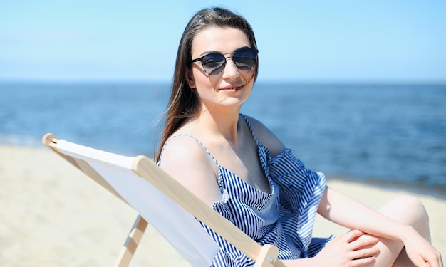 Szczęśliwa młoda brunetka odpoczywa na drewnianym leżaku na plaży oceanu, patrząc w kamerę, uśmiechając się i nosząc modne okulary przeciwsłoneczne. Koncepcja cieszących się wakacji