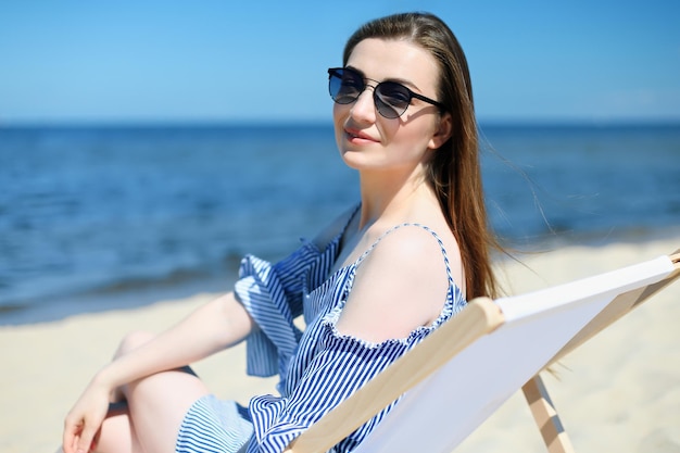 Szczęśliwa młoda brunetka relaksuje się na drewnianym leżaku na plaży oceanu, patrząc na kamerę, uśmiechając się i nosząc modne okulary przeciwsłoneczne. Ciesząca się koncepcja wakacji