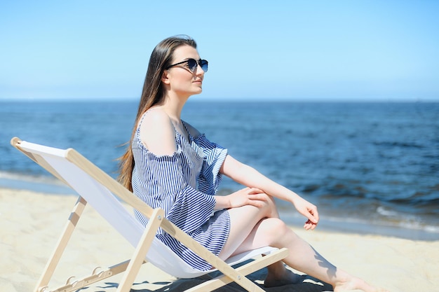 Szczęśliwa młoda brunetka relaksuje się na drewnianym leżaku na plaży oceanu, uśmiechając się i nosząc modne okulary przeciwsłoneczne. Koncepcja cieszących się wakacji