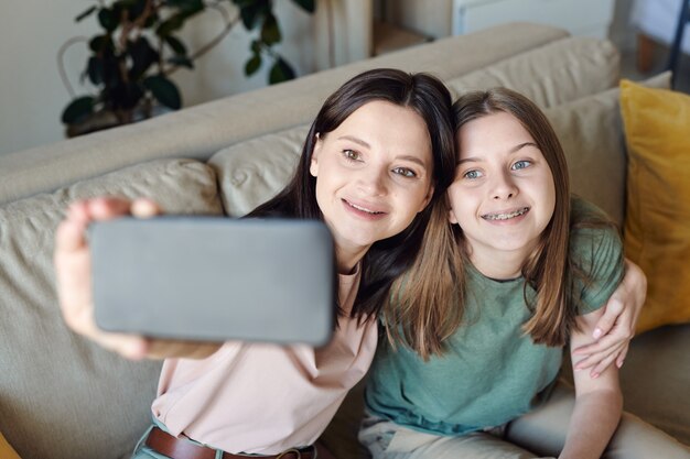 Szczęśliwa młoda brunetka ze smartfonem i jej uśmiechniętą nastoletnią córką robi selfie siedząc na kanapie
