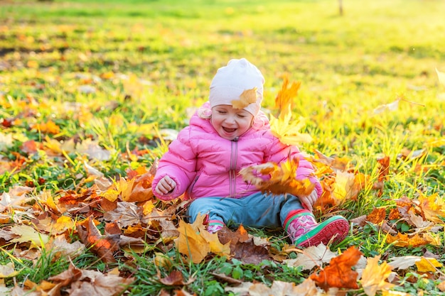 Szczęśliwa młoda dziewczyna gra pod spadającymi żółtymi liśćmi w pięknym jesiennym parku na spacery na łonie natury. Małe dziecko rzuca jesienne pomarańczowe liście klonu. Witam koncepcja jesień.