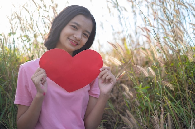 Szczęśliwa młoda dziewczyna trzyma papierowe czerwone serce na łące Azjatycka dziewczyna