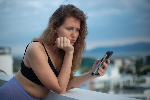 Szczęśliwa młoda dziewczyna używa telefonu na dachu domu, na balkonie, na tle miasta, kobieta robiąca sobie selfie.