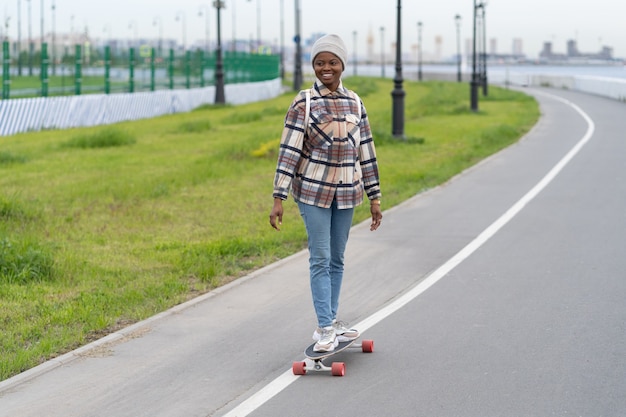 Szczęśliwa młoda kobieta afro na łyżwach na longboardzie, uśmiechnięta, zrelaksowana na deskorolce na świeżym powietrzu w przestrzeni miejskiej