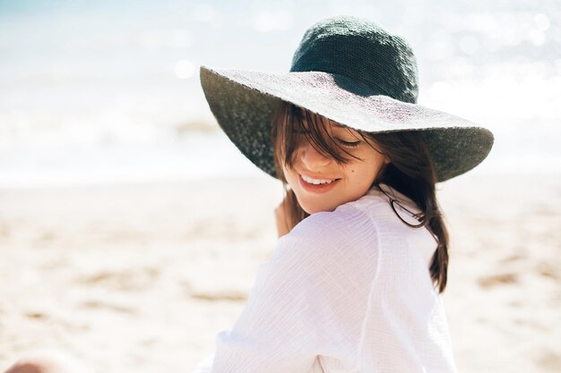 Szczęśliwa młoda kobieta boho w kapeluszu relaksująca się i ciesząca się słonecznym, ciepłym dniem nad oceanem Miejsce na tekst Stylowa dziewczyna hipster siedząca na plaży i opalająca się Letnie wakacje Spokojne emocje