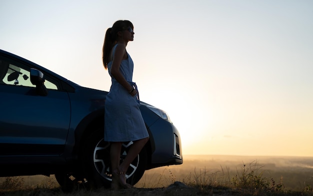 Szczęśliwa młoda kobieta kierowca w niebieskiej sukience, ciesząc się ciepłym letnim wieczorem stojąc obok jej samochodu. Koncepcja podróży i wakacji.