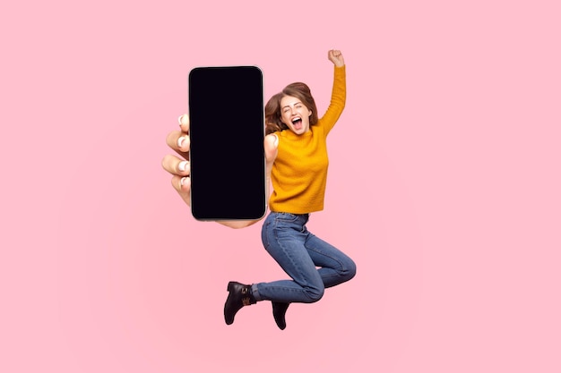 Szczęśliwa młoda kobieta latająca i skacząca w powietrzu i pokazująca duży mobilny pusty ekran dla miejsca kopiowania i powierzchni reklamowej. strzał w studio na białym tle na różowym tle