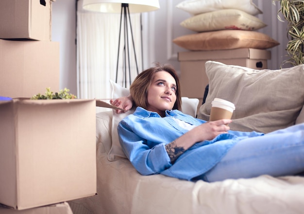 Szczęśliwa młoda kobieta leżąca na kanapie w nowym domu pijąca kawę lub herbatę